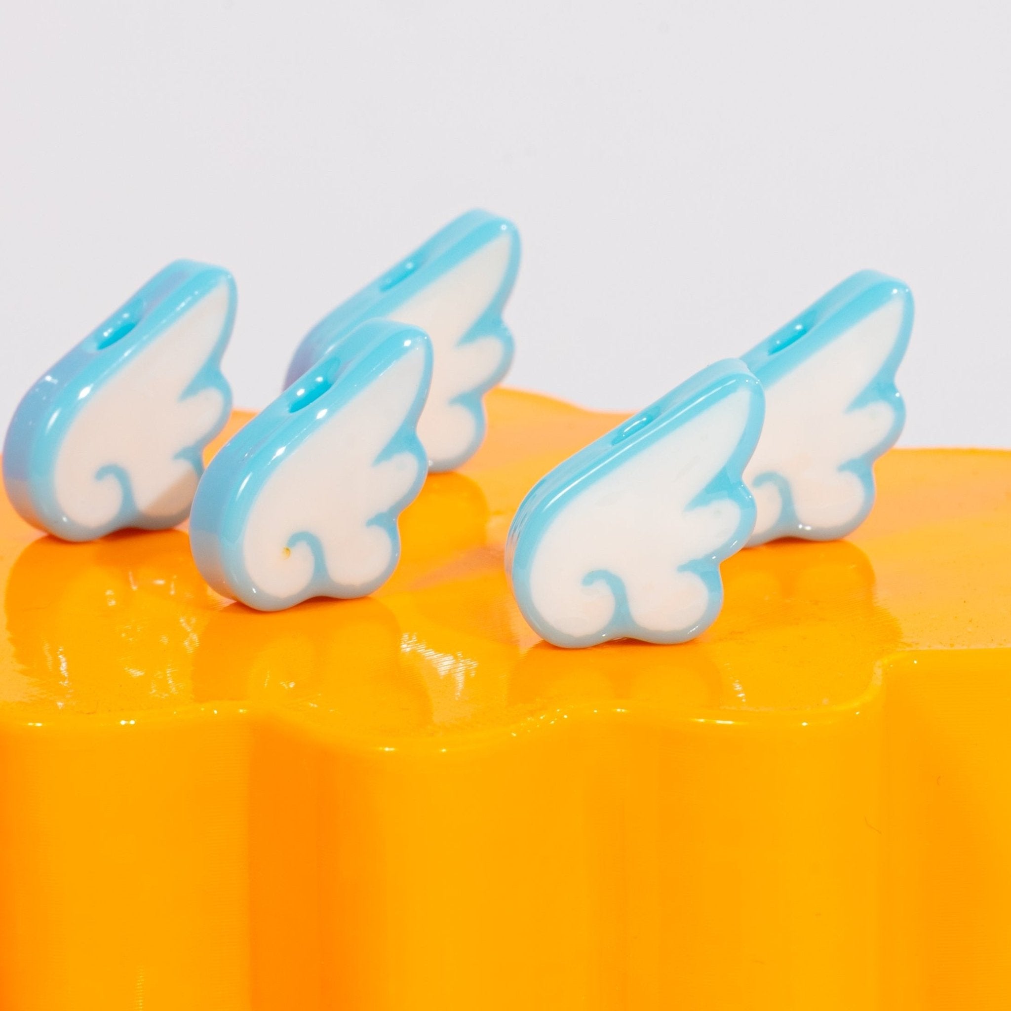 Four white-and-sky-blue angel wing kandi beads lined up on a glossy orange riser