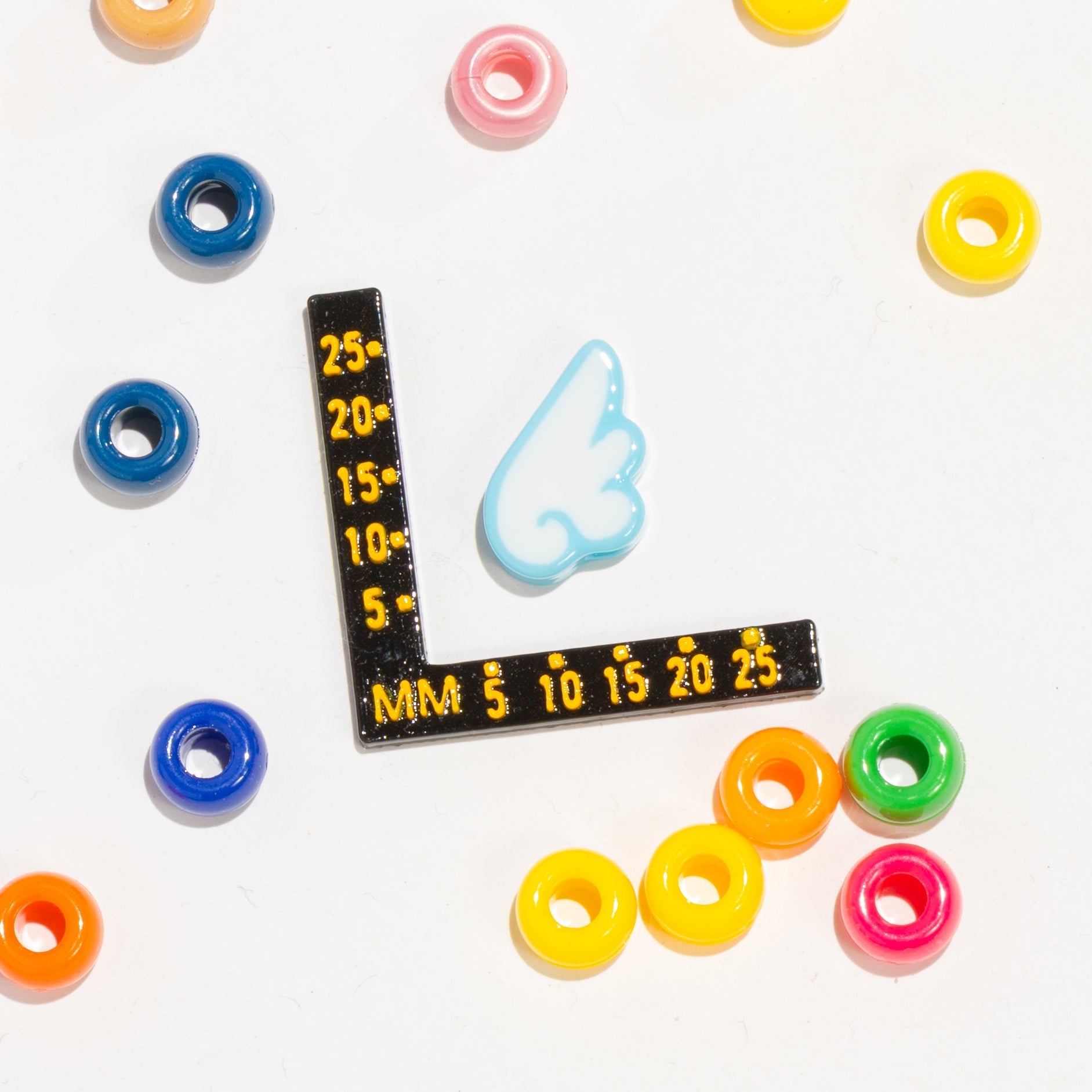 Size guide: a white-and-sky-blue angel wing kandi bead beside a black millimeter ruler, pony beads around