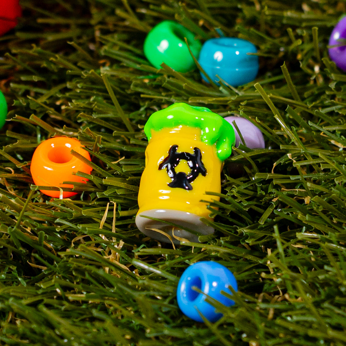 Single biohazard barrel kandi bead in green grass beside colorful beads