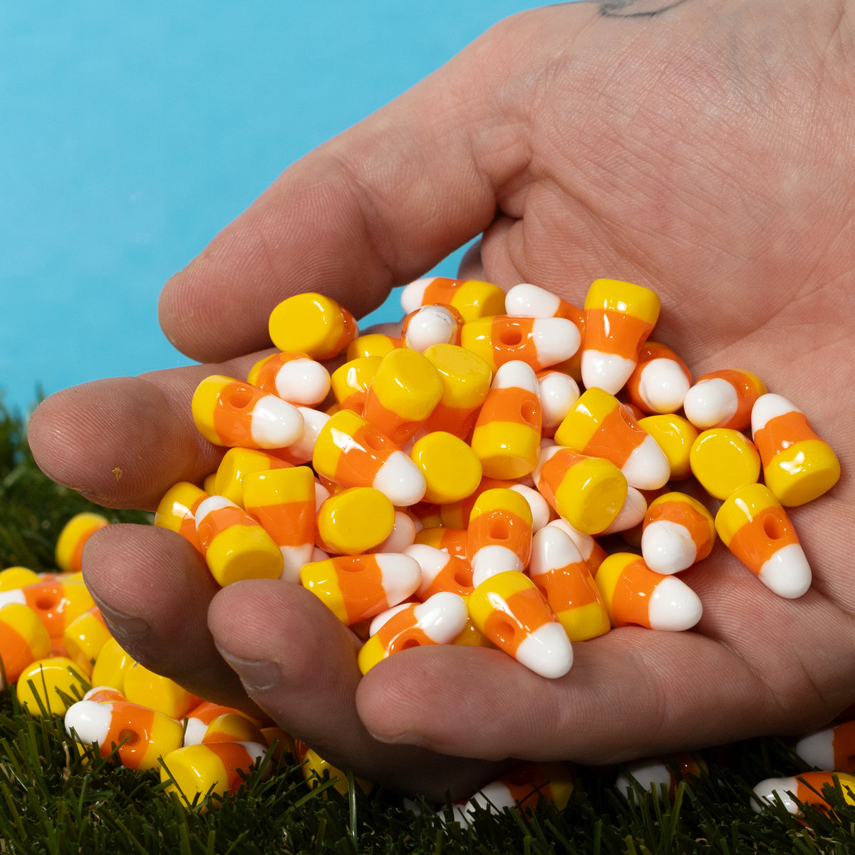 A hand scooping a pile of striped candy corn kandi beads above green grass