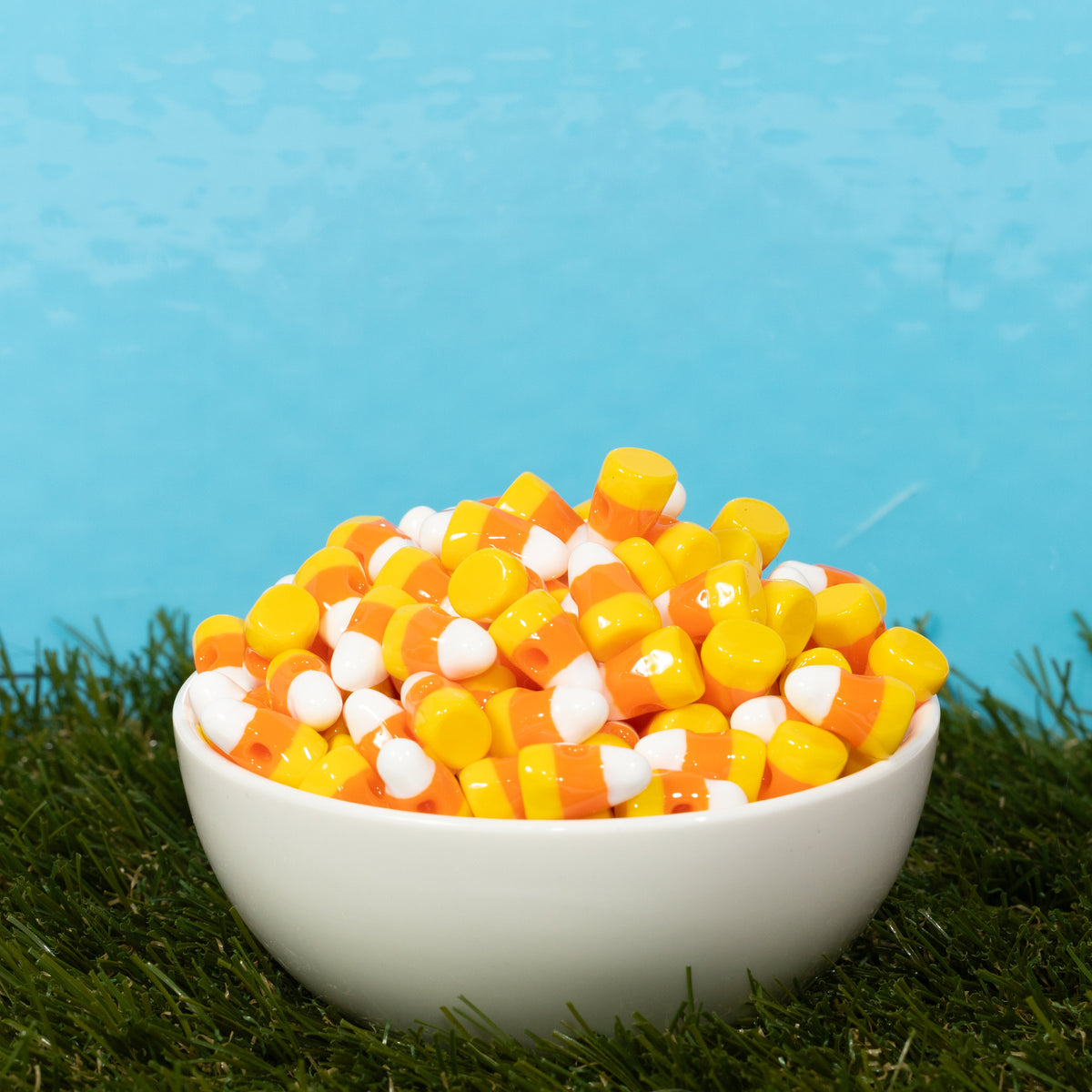 A white bowl overflowing with candy corn kandi beads on grass against blue