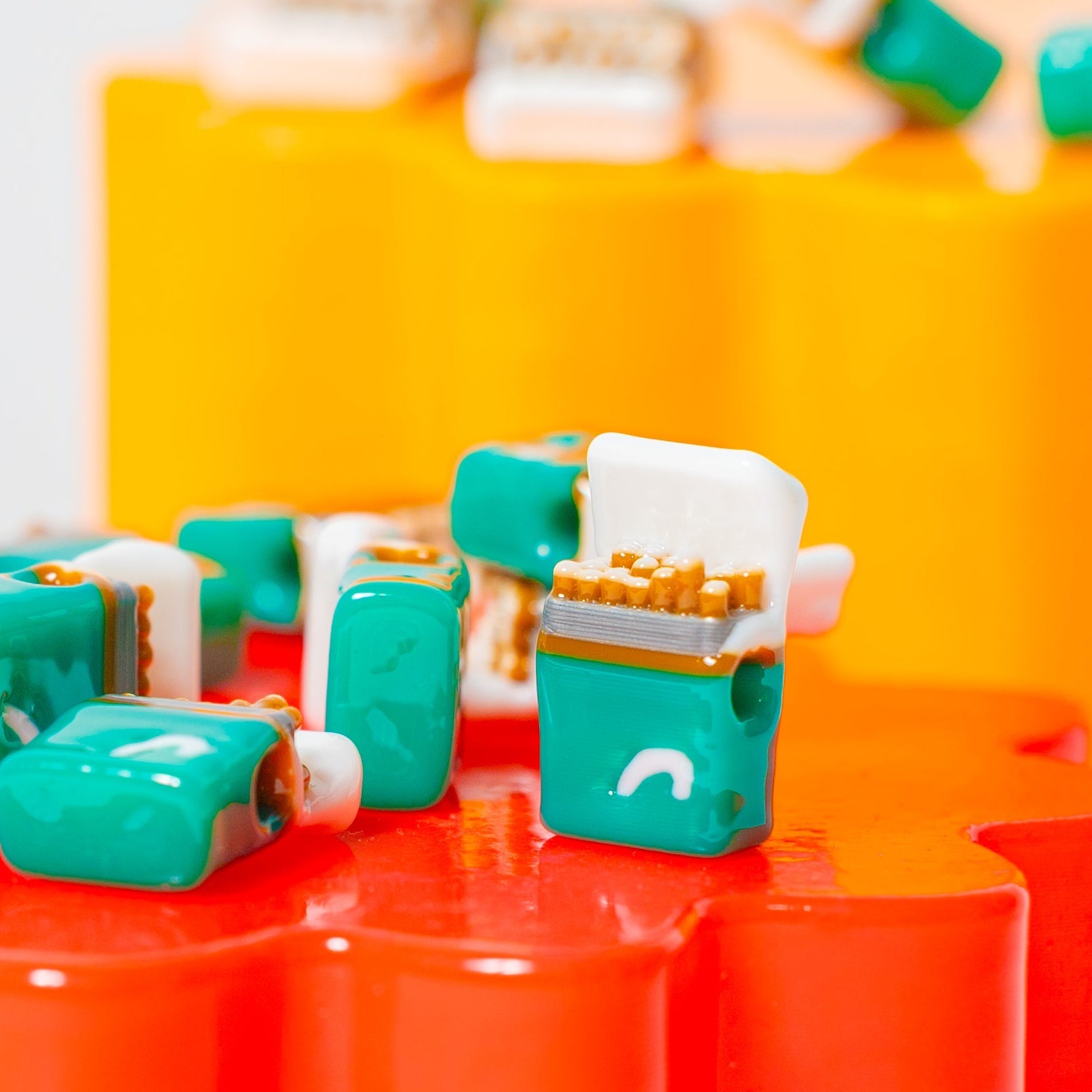 Teal and white cigarette-pack kandi beads on a glossy red riser, orange backdrop behind