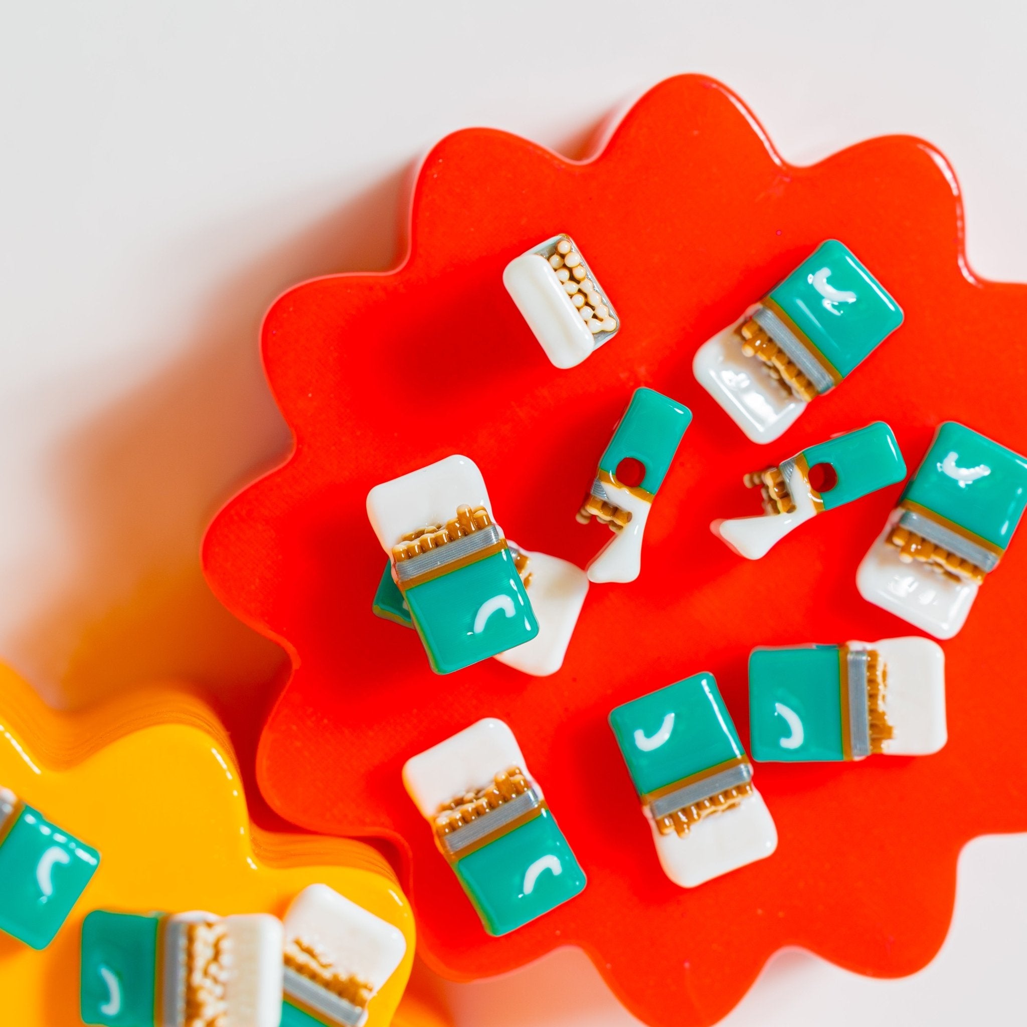 Teal and white cigarette-pack kandi beads scattered on a wavy red plate, one drilled hole showing