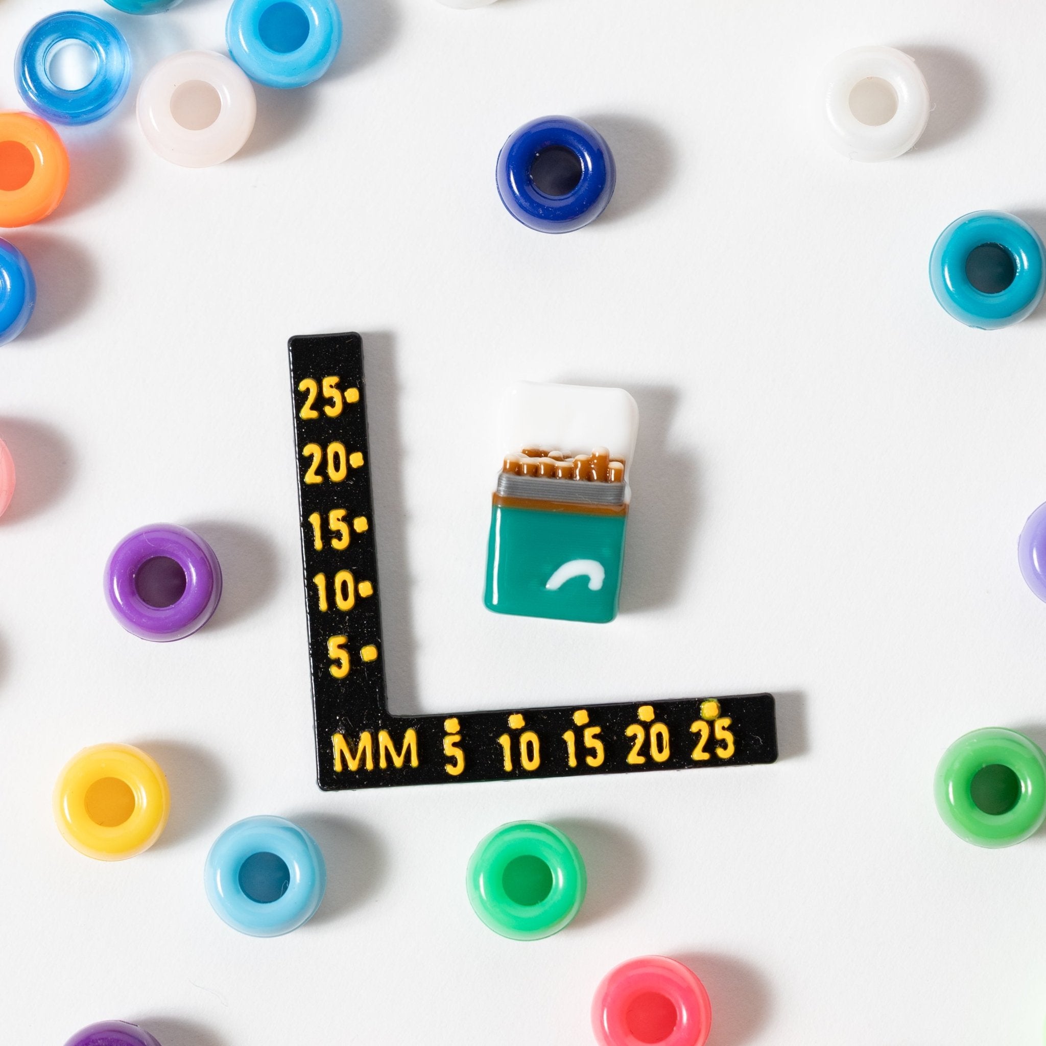 Size guide: a teal cigarette-pack kandi bead beside a millimeter ruler, pastel beads around