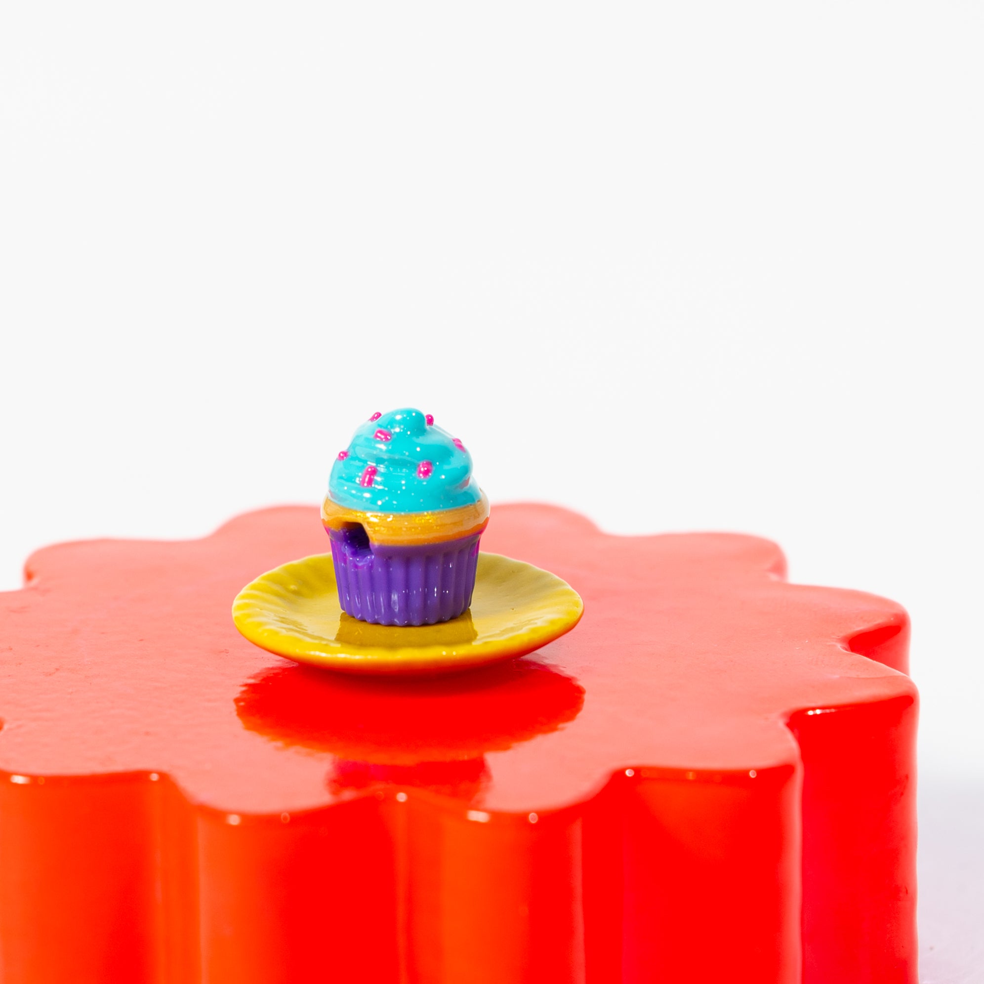 A single cupcake kandi bead on a tiny yellow plate atop a red riser