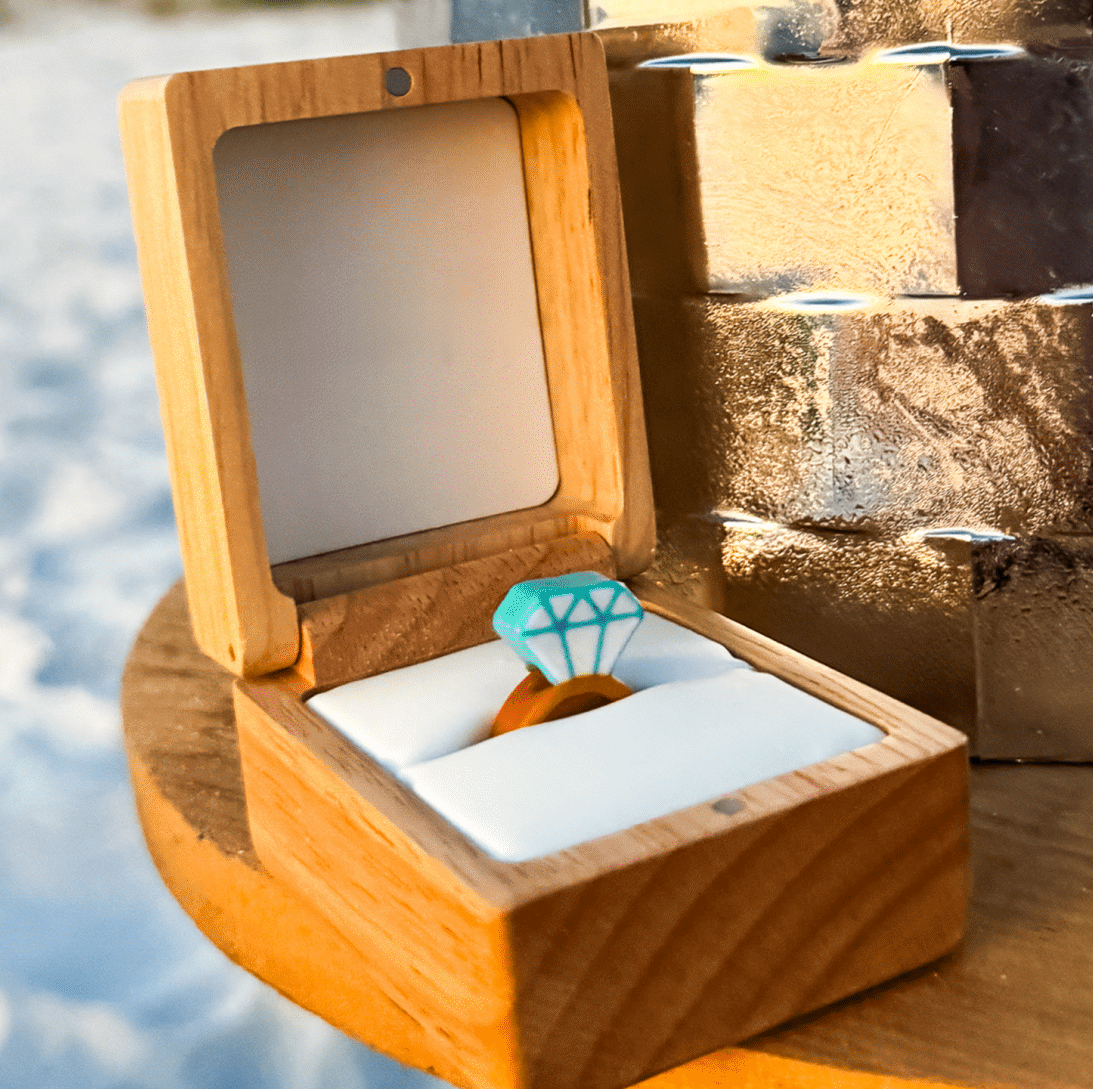 A teal diamond ring bead sits in an open wooden ring box resting on snow in golden light