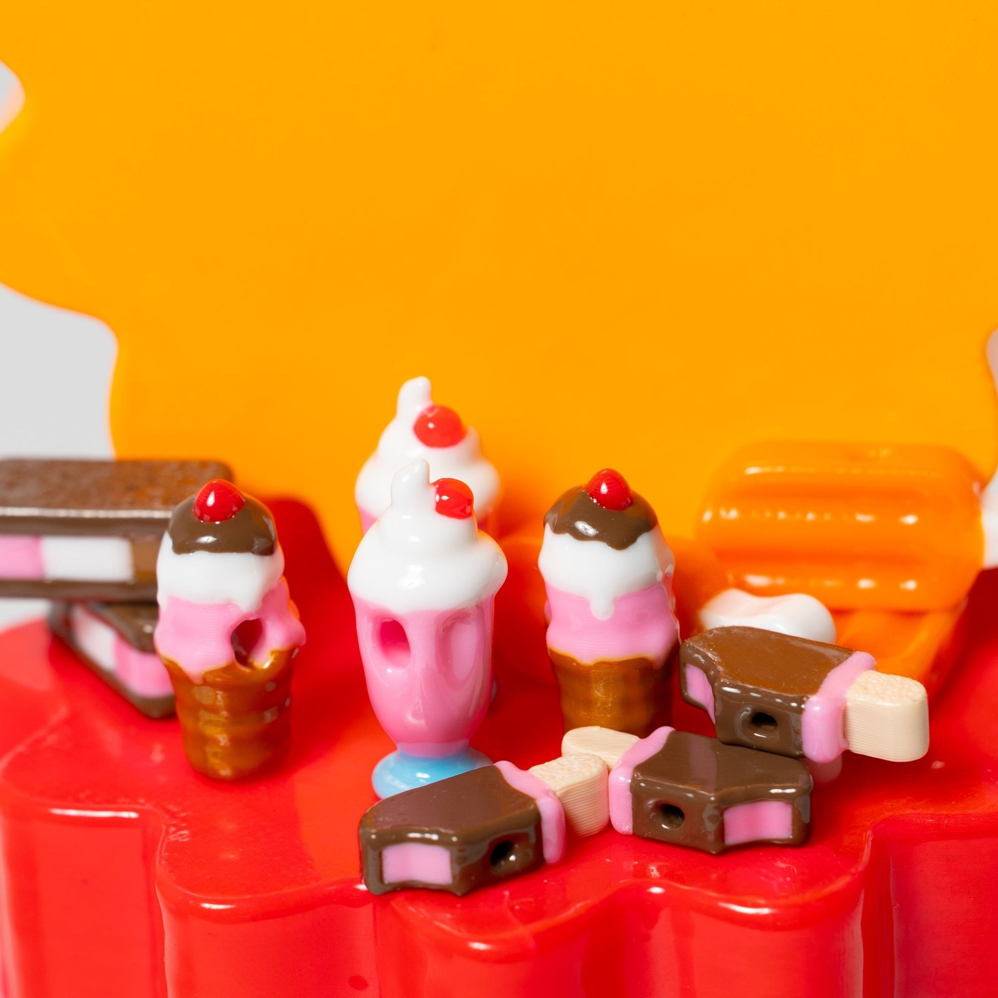 Assorted ice cream kandi beads on red risers: cones, a pink milkshake, sandwiches and chocolate bars, orange backdrop
