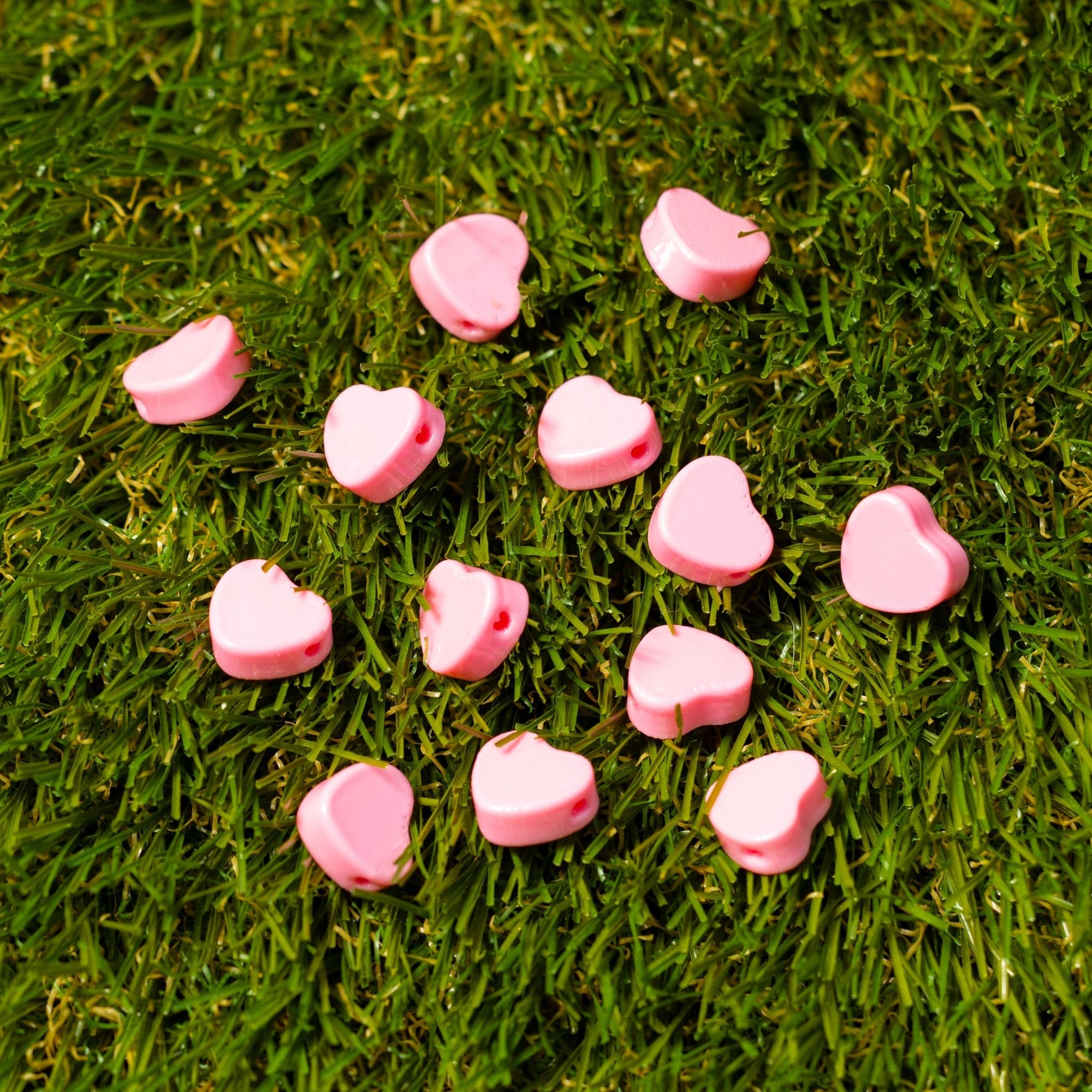 Pink heart kandi beads scattered on green turf, drilled holes catching the light