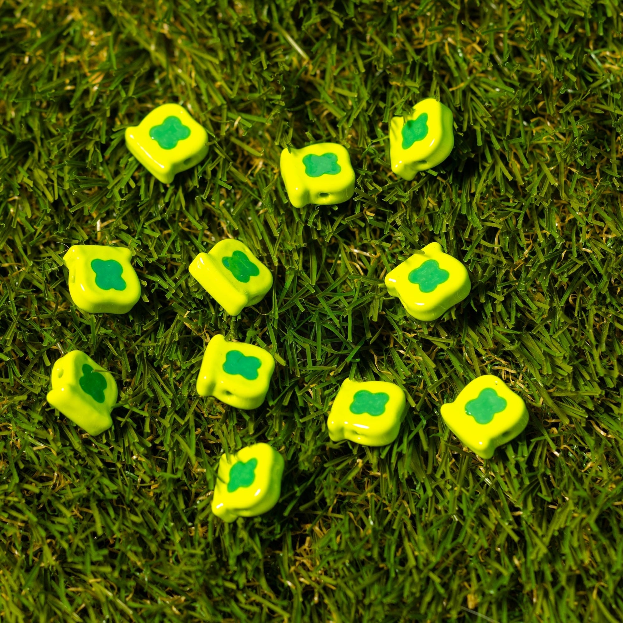 Yellow square kandi beads stamped with a green clover, spread out on artificial grass