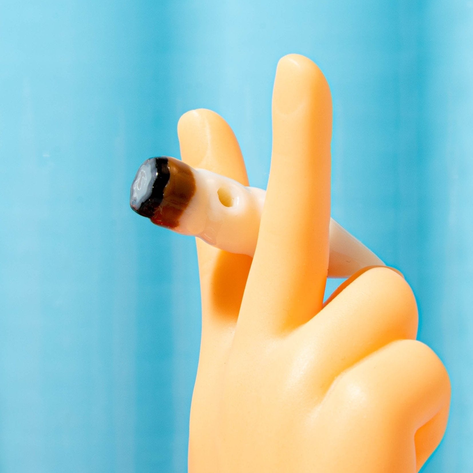 A toy hand holds a lit joint kandi bead between two fingers against a blue backdrop