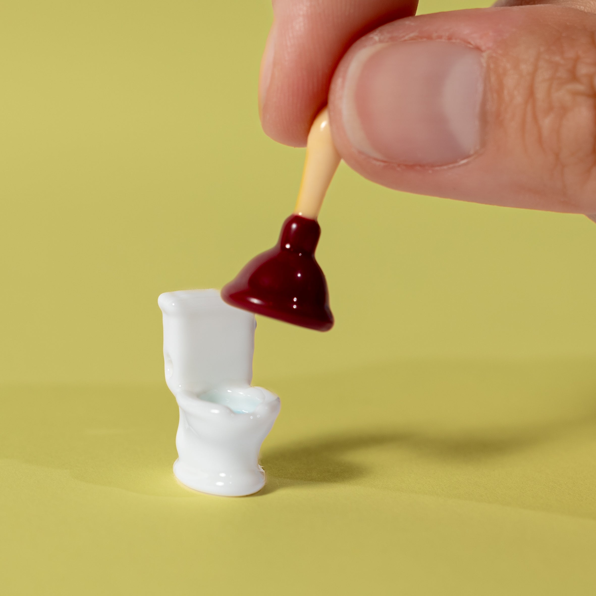 Fingers holding a plunger kandi bead above a tiny white toilet bead on yellow