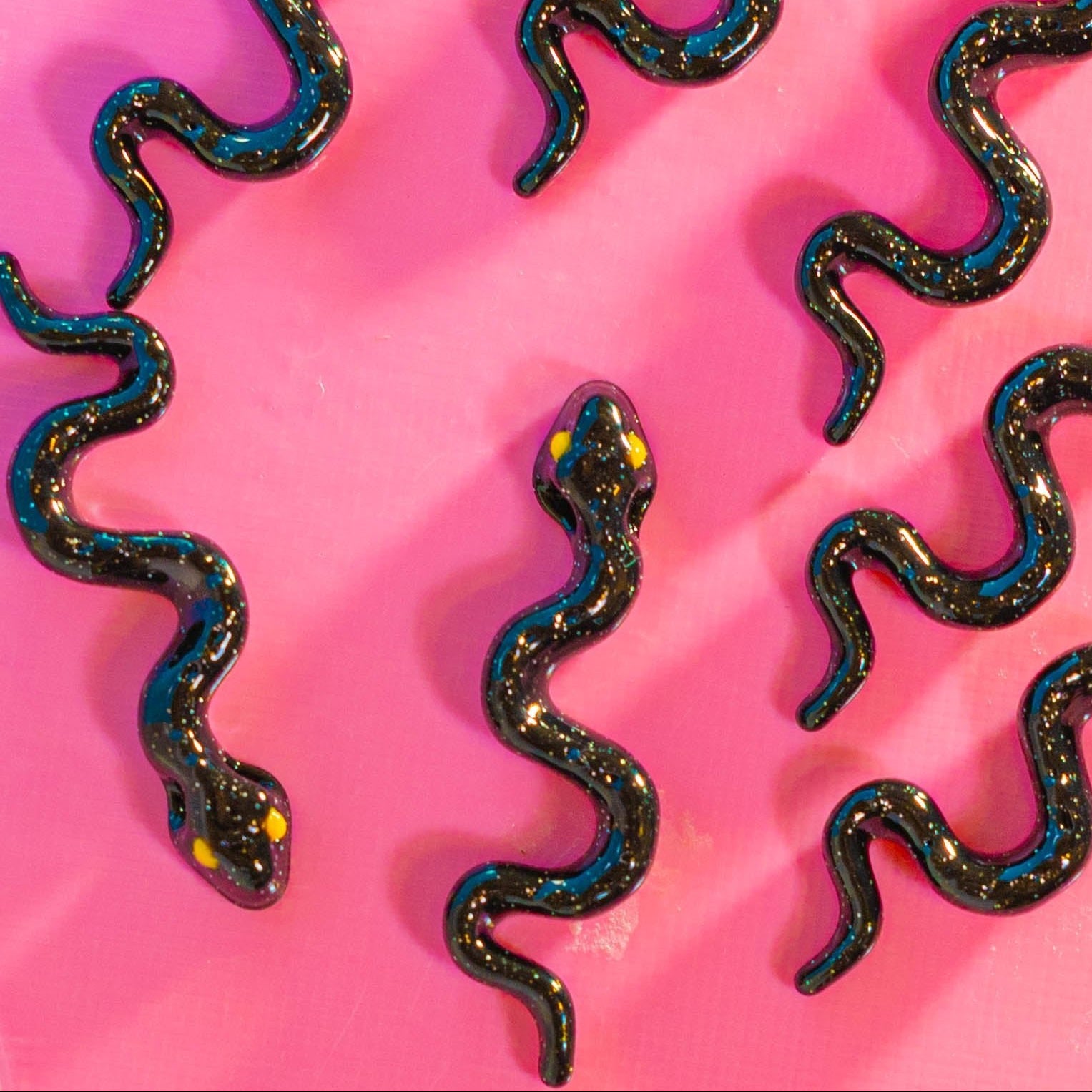 Wavy black glitter snake kandi beads with yellow eyes on a hot pink surface, from above