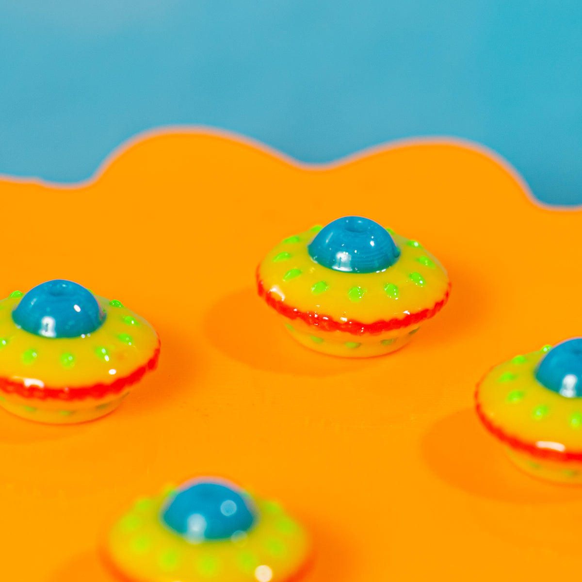 Yellow spaceship kandi beads with red rims and blue domes on a glossy orange riser, blue behind