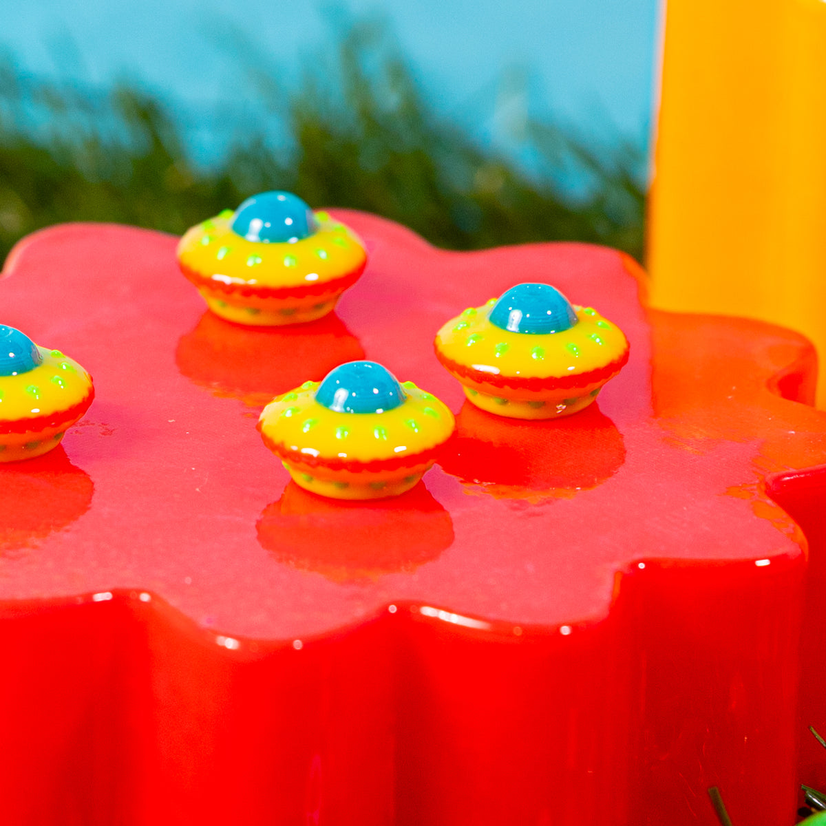 Four spaceship kandi beads on a glossy red riser with green grass and an orange block behind