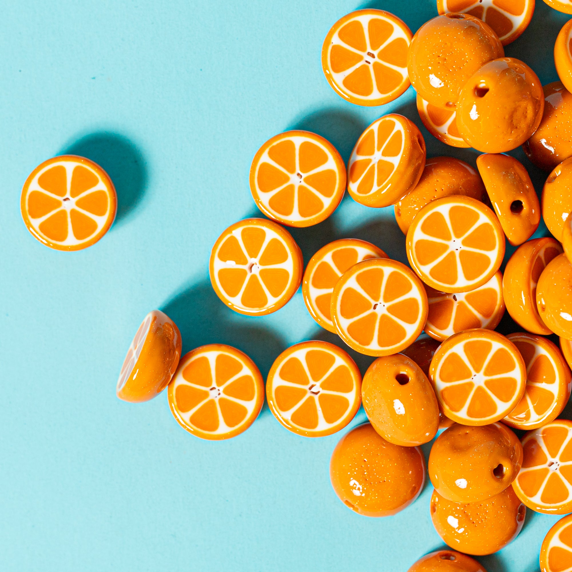 Orange slice kandi beads scattered on teal from above, a few turned to show glossy whole-orange domes