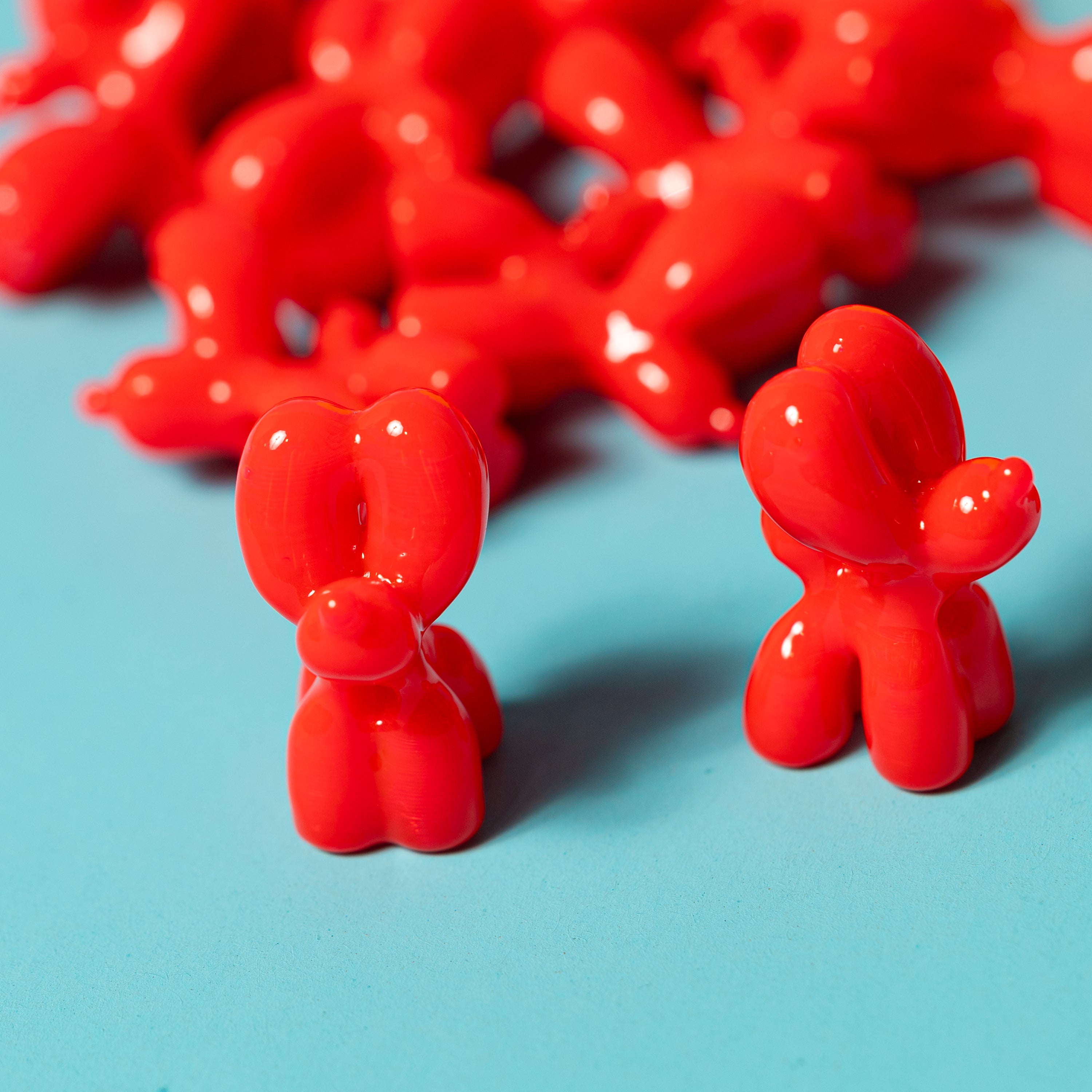 Two glossy red balloon dog kandi beads in front with a pile blurred behind on a teal surface