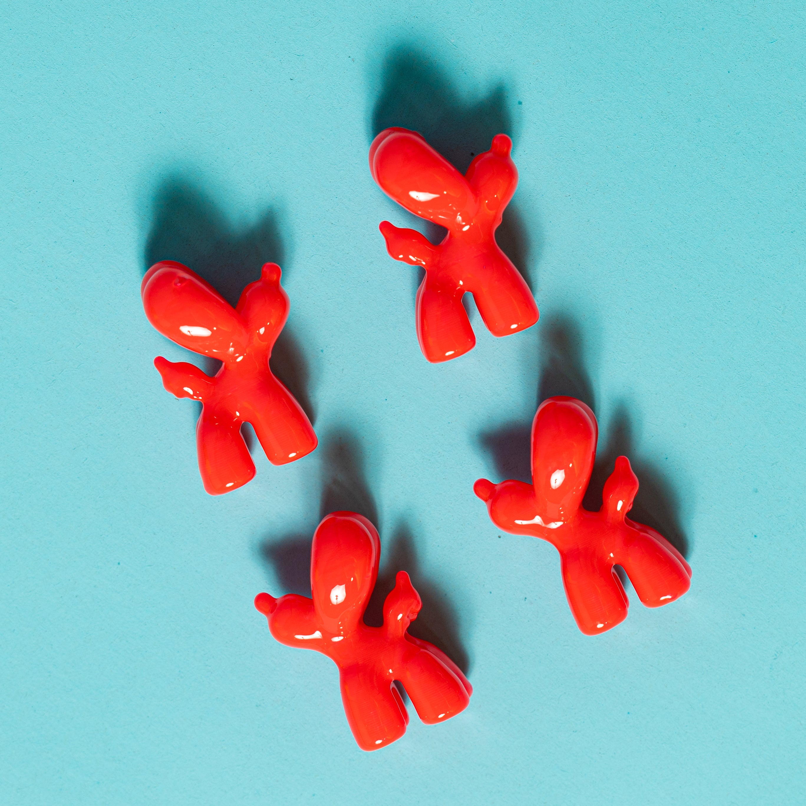 Four red balloon dog kandi beads arranged on teal, casting long shadows, seen from above