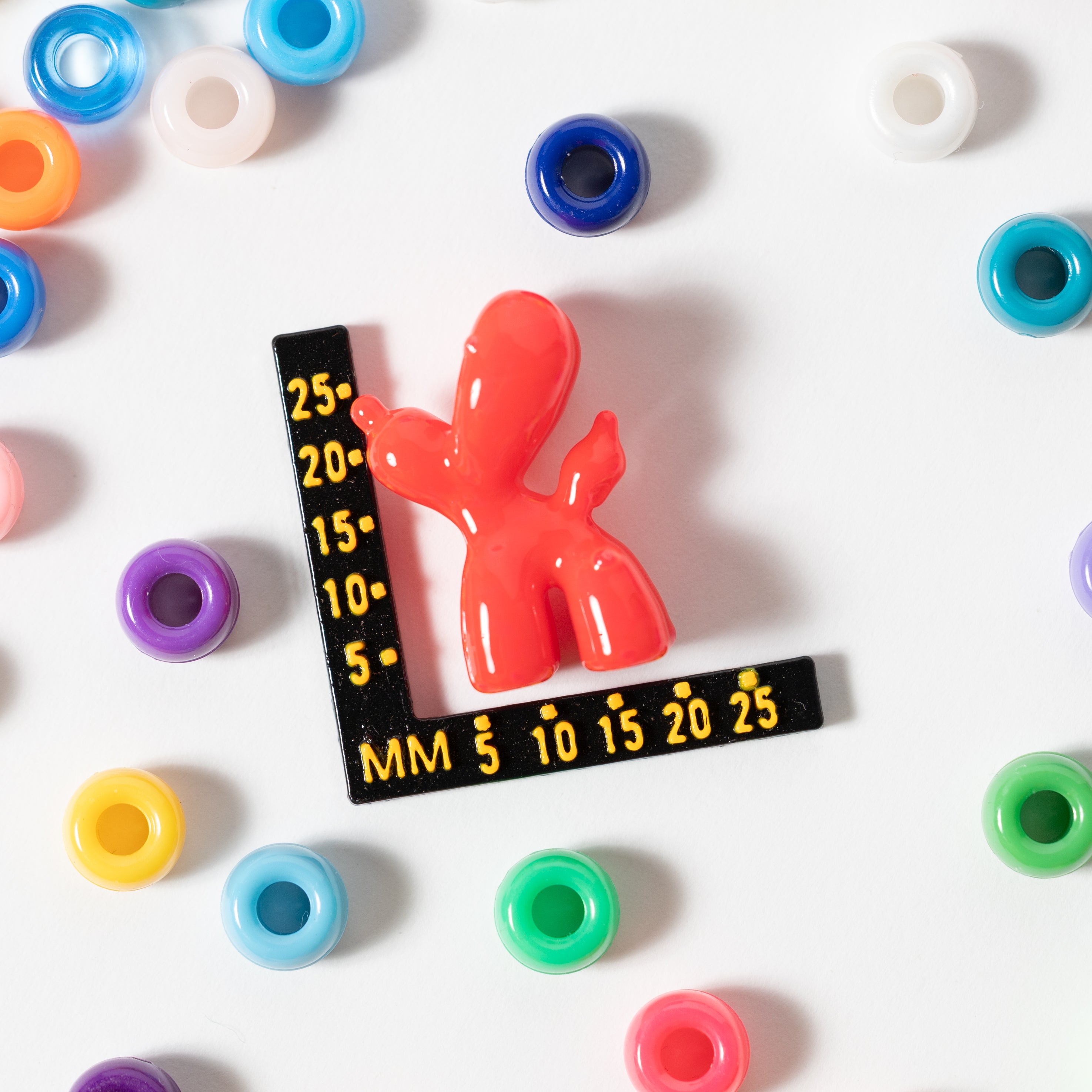Size guide: a red balloon dog kandi bead beside a millimeter ruler, surrounded by rainbow pony beads