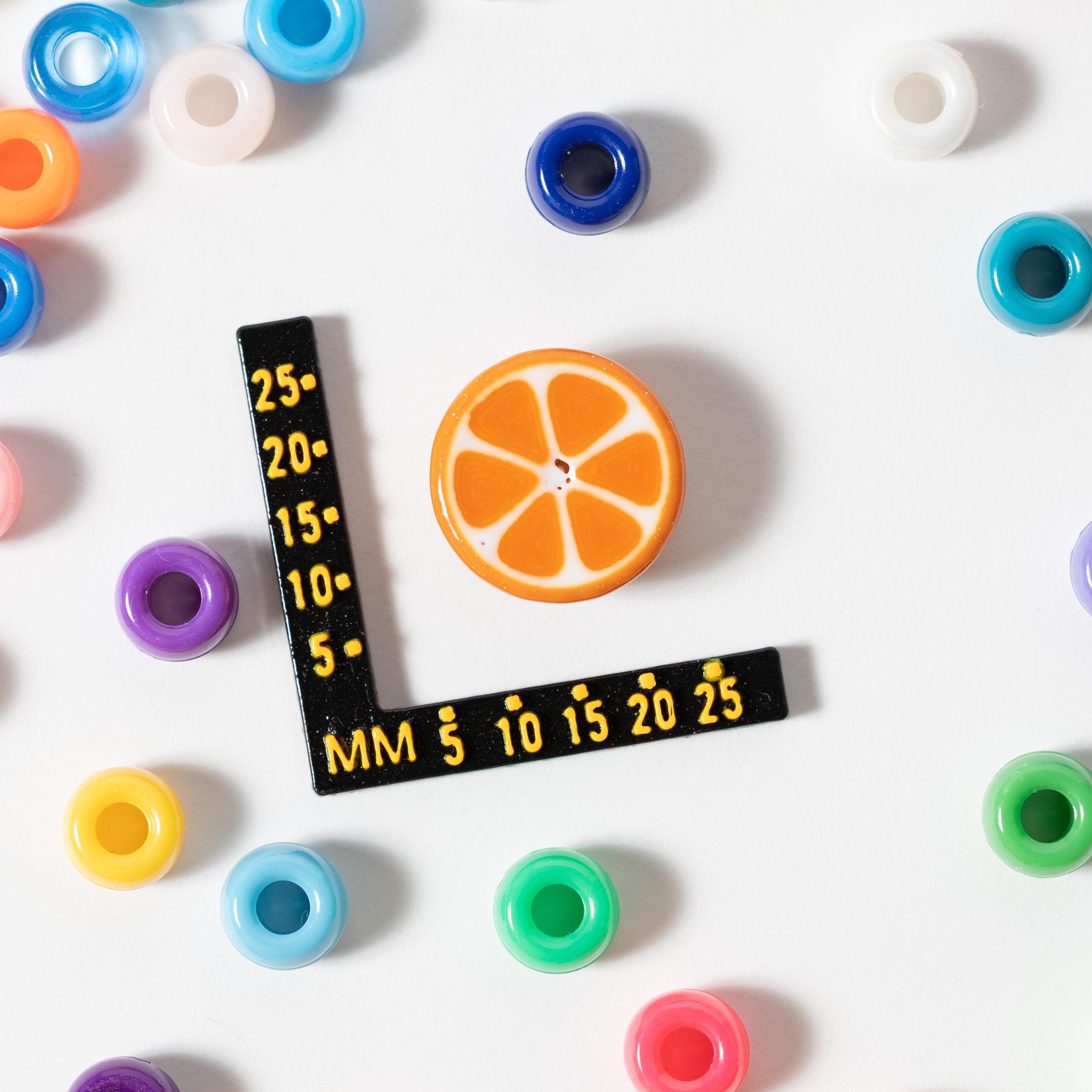 Size guide: an orange slice kandi bead beside a millimeter ruler, surrounded by rainbow pony beads
